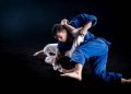 Two judokas in blue and white kimonos wrestling on the ground during a sparring duel. Black background.